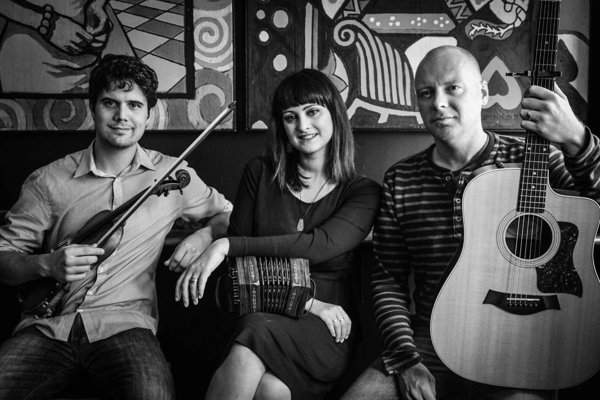 Nathan, Mairéad, and John seated together in a pub with fiddle, concertina, and guitar, colorful paintings on the wall behind them