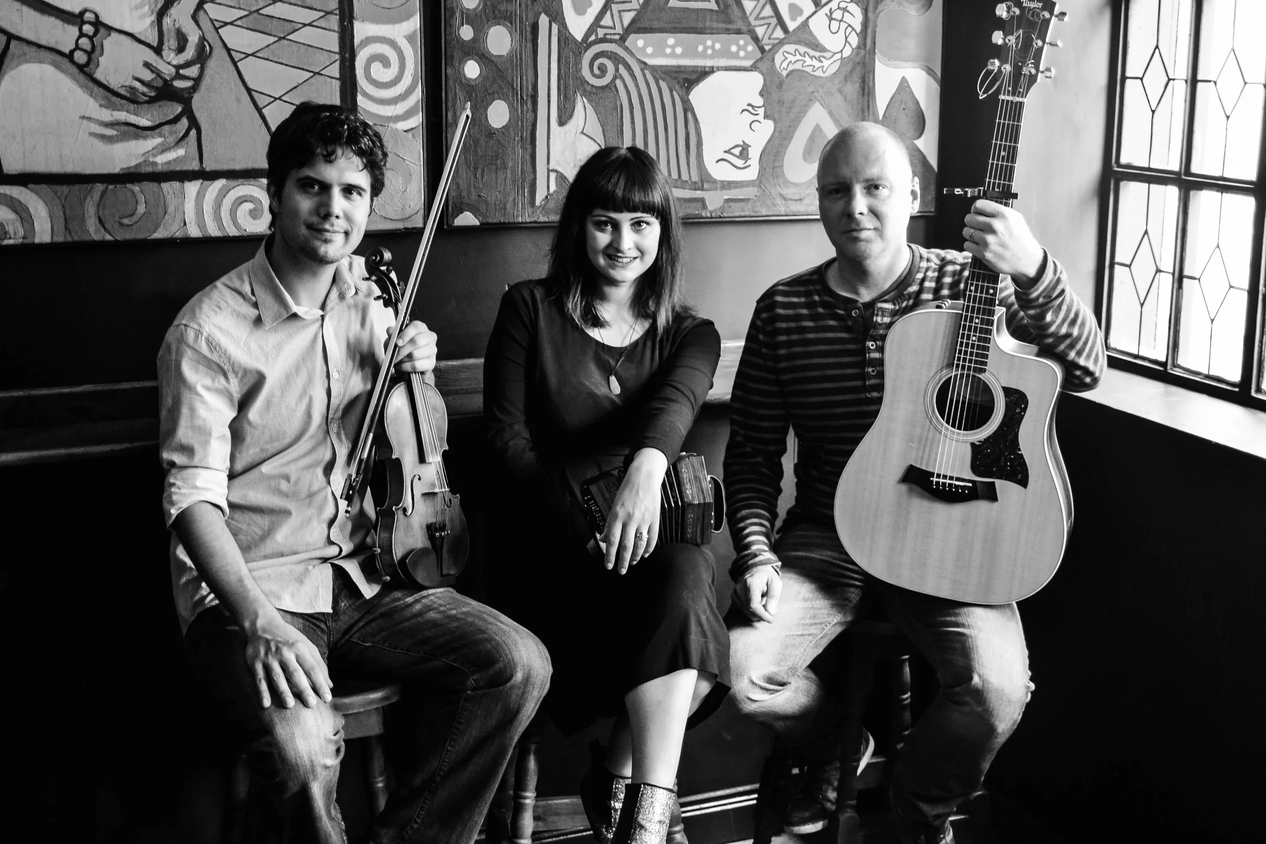 Nathan Gourley, Mairéad Hurley, and John Blake seated with fiddle, concertina, and guitar in a pub with stained glass windows
