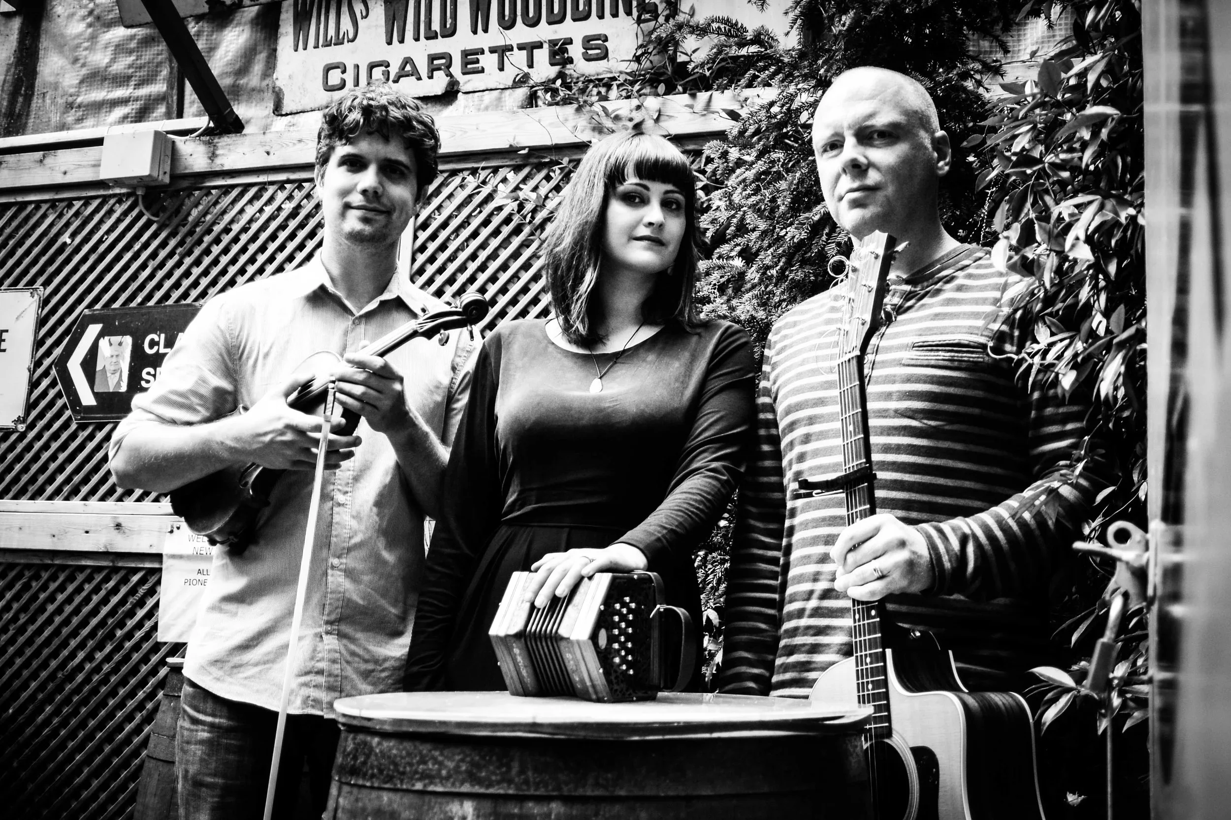 Nathan, Mairéad, and John standing outdoors by a wooden barrel with their instruments, vintage signage in the background